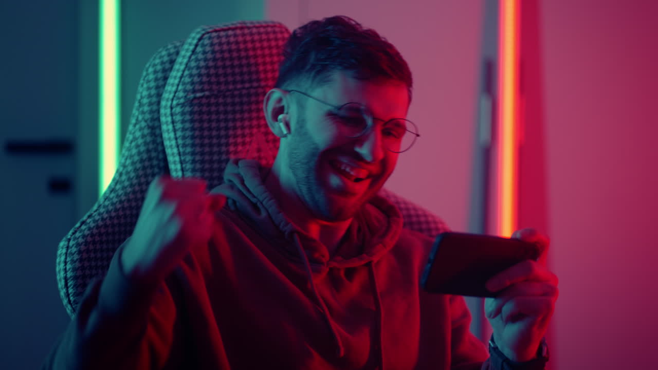 joven jugando videojuego por teléfono inteligente regocijándose ganar jugador emocional ganando en juego de tiro retrato de jugador de videojuego en casa en la noche