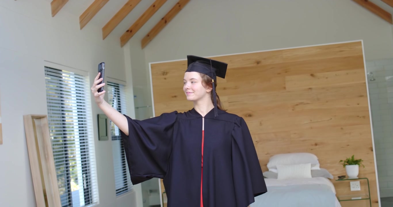 Laureata con tocco e toga che alza il telefono per un selfie, legge e risponde a messaggi in camera da letto.