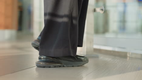 close up leg view of person standing near glass railing in indoor modern setting with blurred background light reflecting off polished floor and casual black sneakers paired with loose black trousers