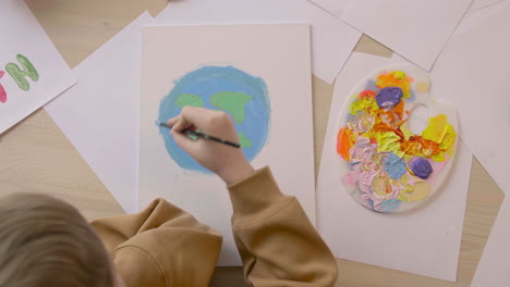 Top-View-Of-Blond-Kid-Painting-A-Earth-On-A-Paper-Sitting-At-A-Table-Where-Are-Signs-With-Environmental-Quotes