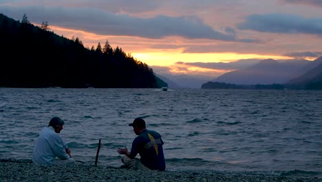 Two-people-talking-while-boat-goes-by-with-vibrant-sunset-on-a-lake-with-mountains-and-clouds-in-background