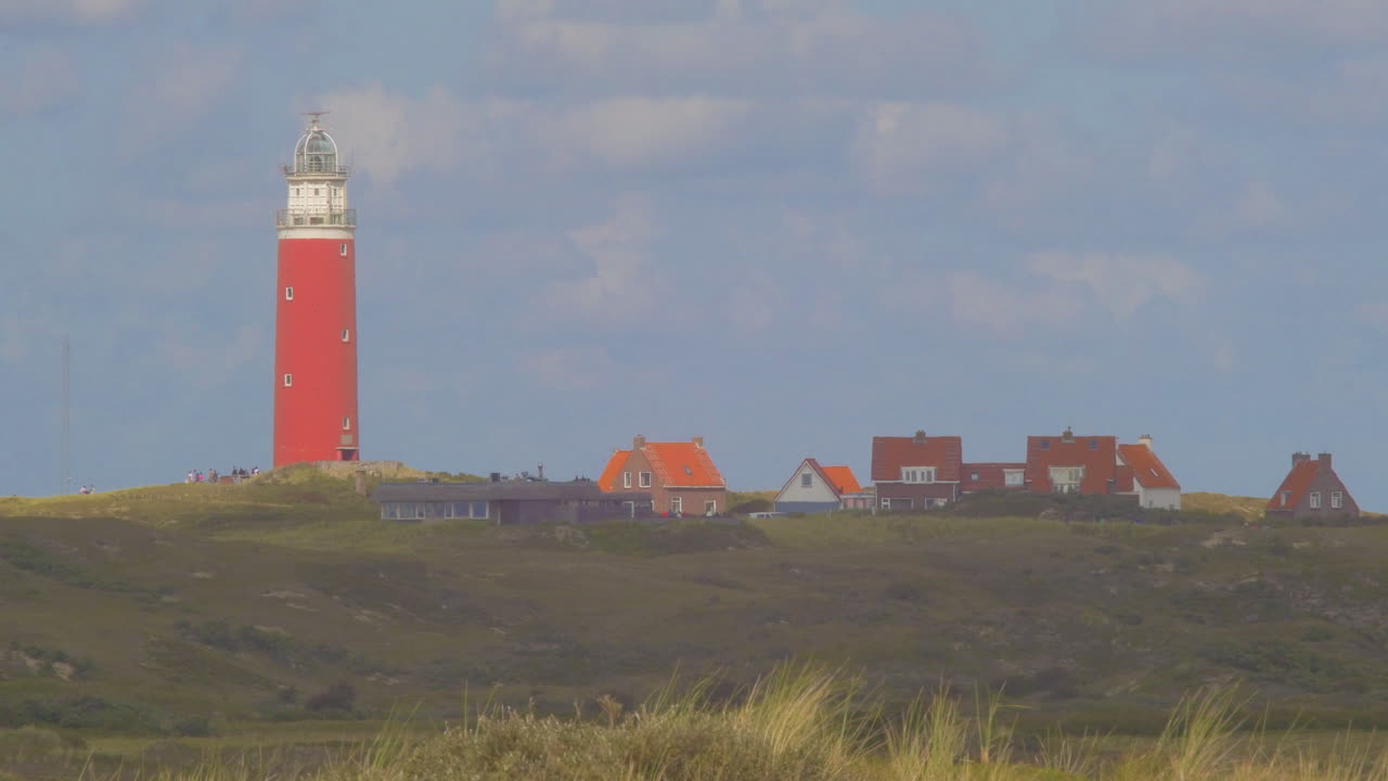 Steady Tele shot of Waving Marram grass in the foreground. In the distance we see a lighthouse in the dunes together with some builings. The sky is blue with seagulls and beautiful clouds passing by.