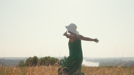 woman in green dress and white hat stretches arms joyfully while turning around in golden grassy field under bright sky with river landscape in distance
