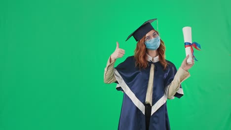 educación a distancia, graduada femenina con sombrero académico y manto con máscara médica en la cara apunta al diploma, luego el dedo hacia arriba y la mano al lado lugar para la inscripción mientras está de pie sobre fondo verde, llave cromada