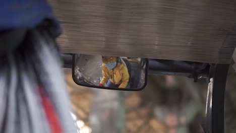 Inside-the-warehouse-electric-car.-Female-worker-in-hard-hat-and-yellow-jacket-and-mask-sitting-in-machine,-looking-to-the-backview-mirror.-Woman-driver,-sorting-stacks-of-pressed-carton