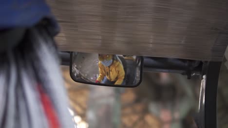 dentro del almacén coche eléctrico. trabajadora en casco y chaqueta amarilla y máscara sentada en la máquina, mirando al espejo retrovisor. mujer conductor, clasificando pilas de cartón prensado