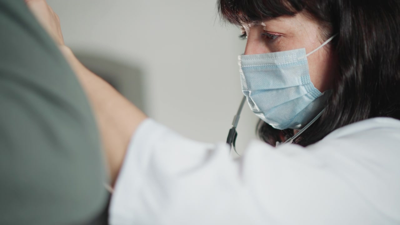 Clínico enmascarado escucha la parte baja de la espalda con estetoscopio durante una consulta cercana, enfoque profesional con mascarilla protectora, iluminación clínica tranquila, técnica cuidadosa y tranquilidad del paciente.