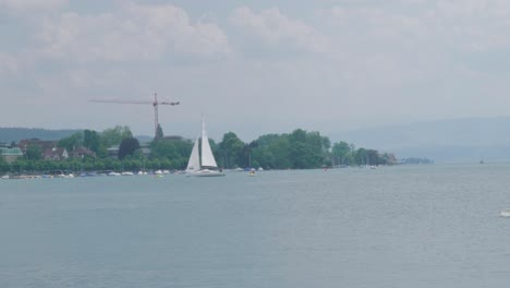 scenic drone footage of a sailing boat with a swiss flag sailing on a beautiful lake at the foot of the alps, not far from a village located on the coast