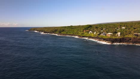 Afternoon-drone-shot-reveals-Pahoa-houses-perched-on-cliffs-while-Pacific-waves-crash-below,-under-clear-Hawaiian-skies