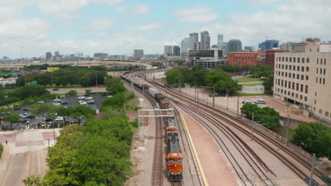 Vista-Aérea-Del-Tren-De-Carga-Que-Recorre-Lentamente-La-Estación-Ebj-Union.-Drone-Volador-Hacia-Atrás-Siguiendo-El-Corredor-Ferroviario.-Horizonte-Con-Rascacielos-De-Fondo.-Dallas,-Texas,-Nosotros