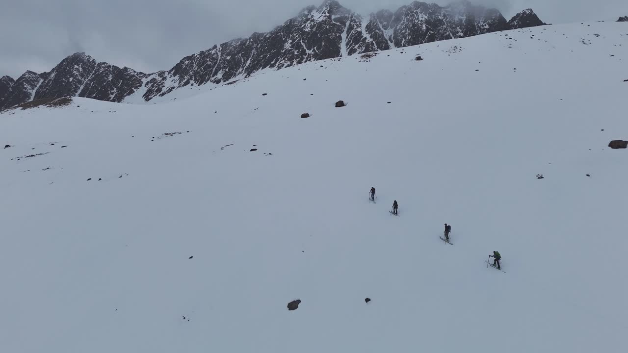 A group of ski tourers makes their way up a wide snow-covered slope below dramatic alpine peaks, capturing the solitude, effort, and beauty of a remote mountain environment. Location: Alps