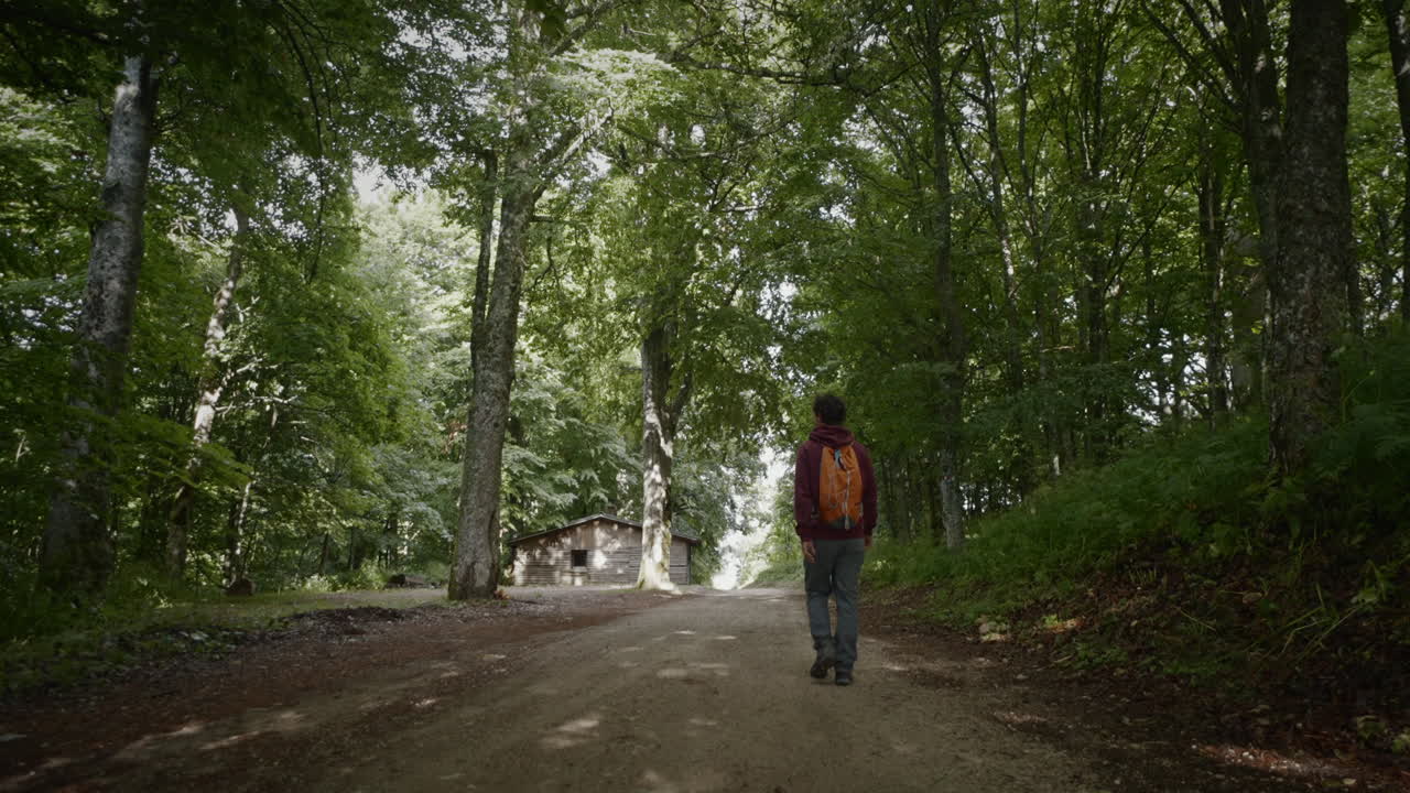 Gimbal tracking a young hiker with a orange backpack and gurgundy red hoody walking on a forest path walking towards the forest cabin