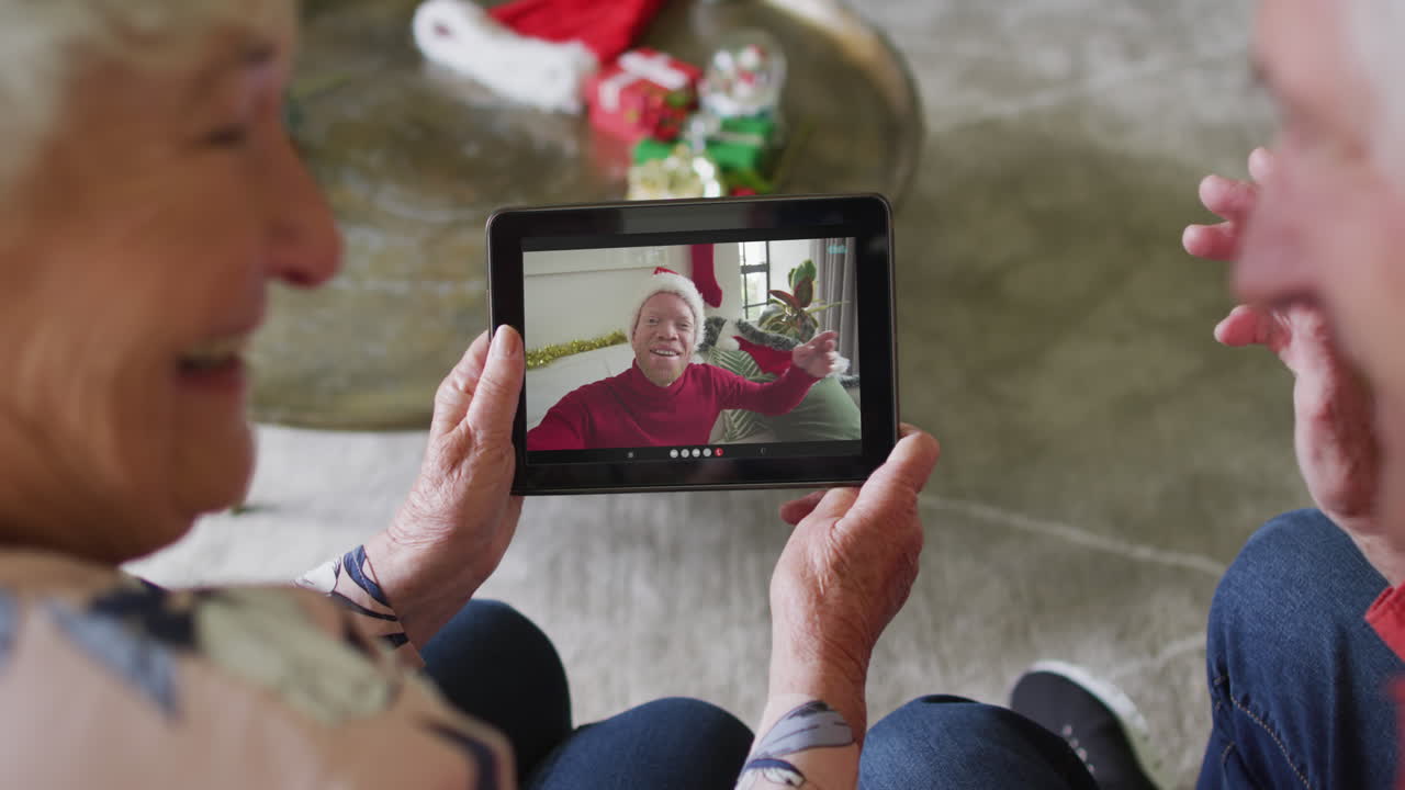 Coppia anziana caucasica sorridente che usa un tablet per una videochiamata di Natale con un uomo sullo schermo.