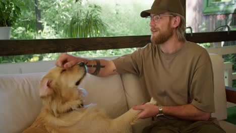 A-blond-guy-with-a-beard-and-glasses-in-a-light-brown-T-shirt-and-cap-is-training-and-teaching-a-new-team-a-large-light-colored-dog-on-a-sofa-in-a-gazebo-in-nature.-The-dog-receives-a-reward-in-the-form-of-a-treat-for-the-executed-command