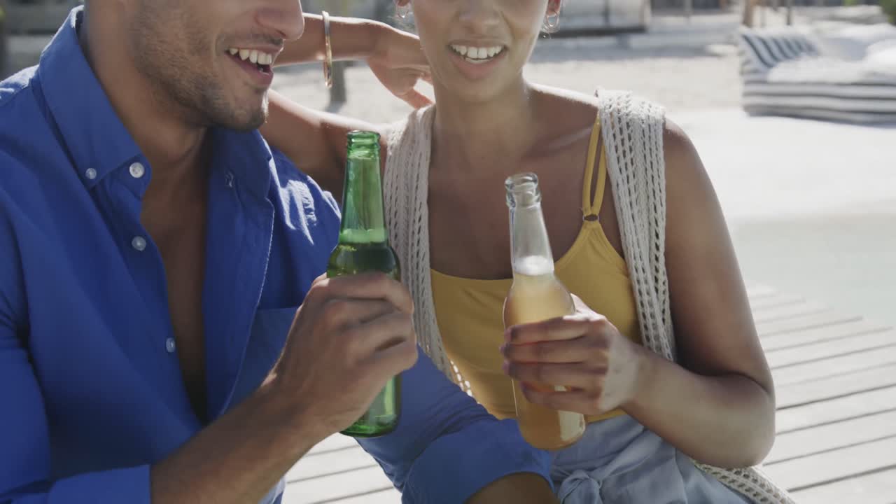 feliz pareja diversa sonriendo y bebiendo un brindis con cervezas en la cubierta del sol de la playa, en cámara lenta