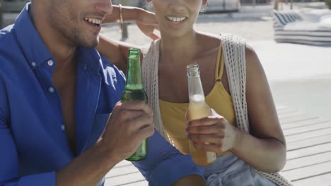 Happy-diverse-couple-smiling-and-drinking-a-toast-with-beers-on-beach-sun-deck,-in-slow-motion