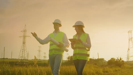 dos electricistas trabajan juntos de pie en el campo cerca de la línea de transmisión de electricidad en cascos. dos electricistas trabajan juntos de pie en el campo cerca con torres de transmisión de energía. combustible ecológico
