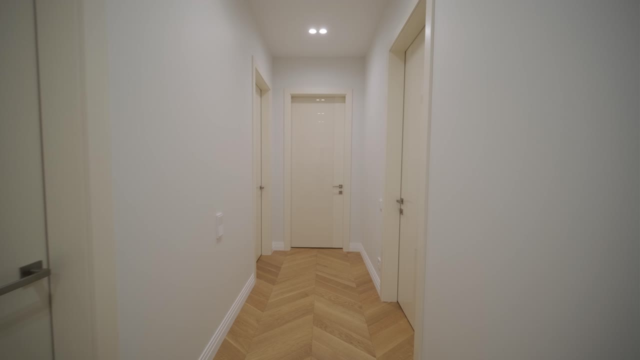 un corredor brillante y estrecho con un piso de madera con patrón de espina de pescado y paredes de tonos neutrales, con múltiples puertas y iluminación moderna encajada, captura un interior limpio y contemporáneo.