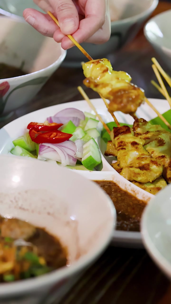 Close-up tangan mencelupkan sate daging panggang ke dalam saus kacang dengan hiasan salad samping