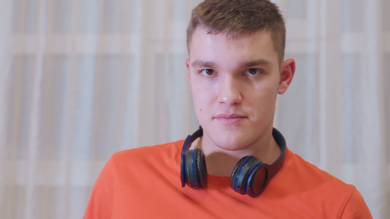 Profile view of young man in orange shirt with headphones around neck giving warm expression while sitting indoors against curtain background