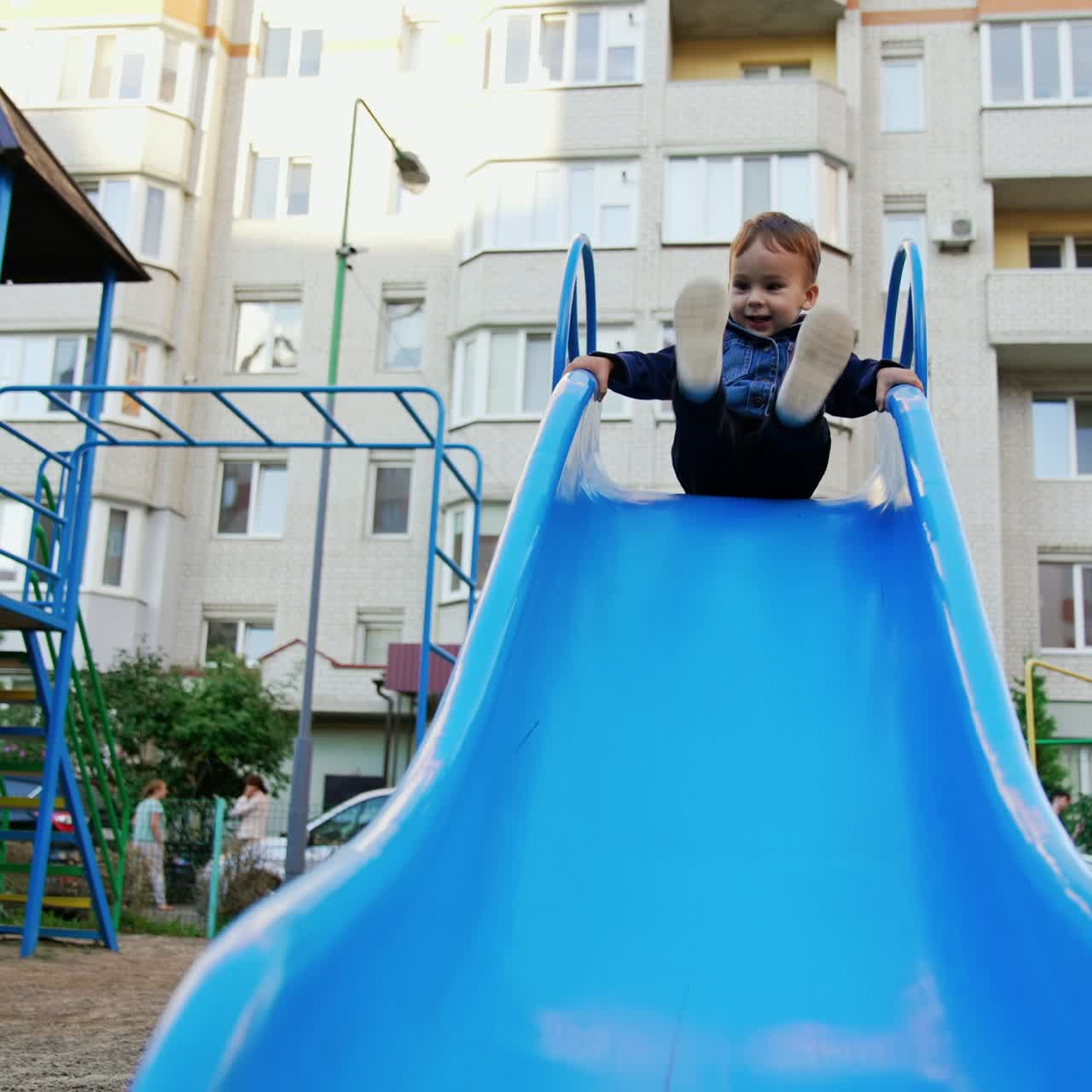 Adorable toddler boy wearing jeans jacket sits on the top of a slide. Kid waves his feet and then goes down. Low angle view