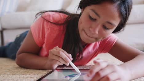 Teenage-girl-lying-on-the-floor-in-the-living-room-using-a-tablet-computer-and-stylus,-close-up