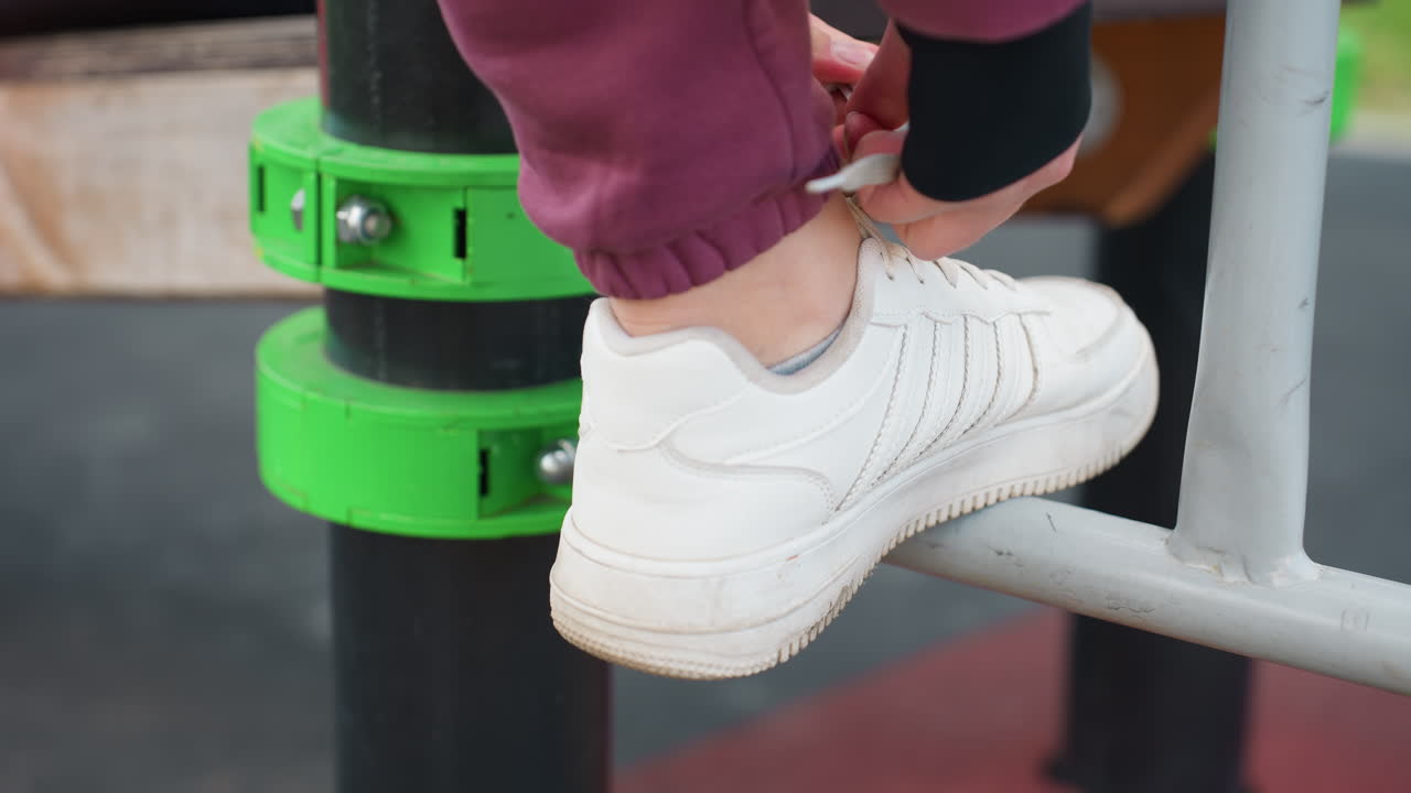 High angle sunlit view of active woman tying sneaker laces on iron workout bar as foot rests on metal rail before starting urban outdoor fitness session on rubber mat near modern apartment complex