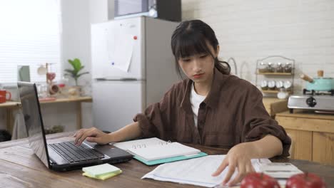 portrait of a concentrated chinese female is recording data in notebook while filing tax return online on the laptop in the dining room at home during daytime.