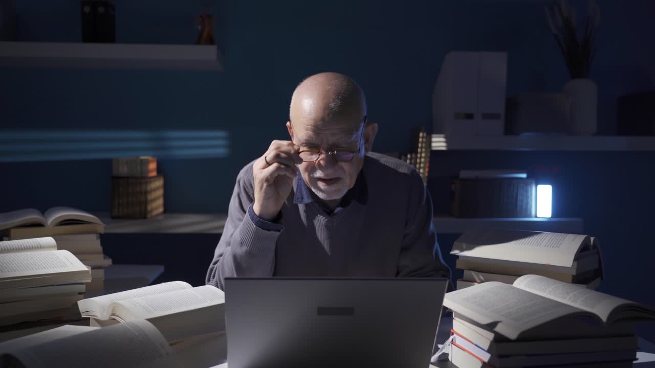 Thoughtful elderly researcher using laptop and browsing scientific books in his office.