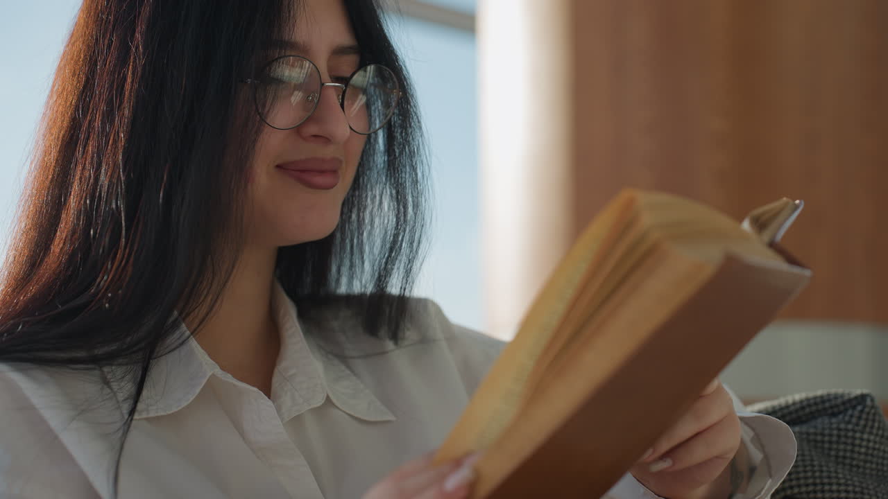 Smiling lady with long black hair and glasses enjoying novel in brightly lit space, dressed in white shirt, resting on wooden rail with blurred urban backdrop and warm natural light