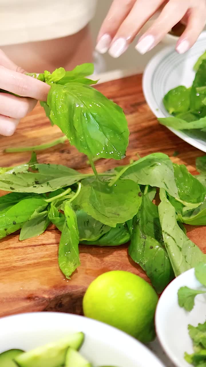Primo piano di mani che staccano foglie di basilico fresco su un tagliere di legno con lime e cetriolo