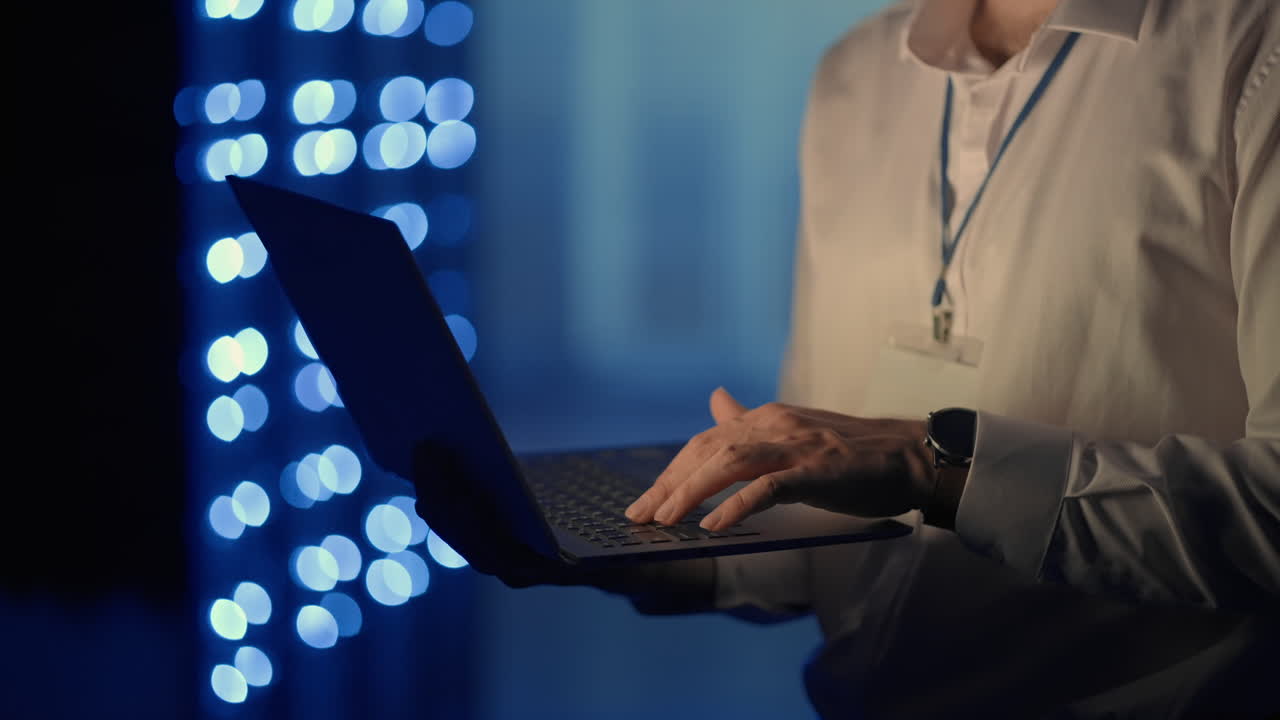 Male network engineer doing a system check standing in the server room with his laptop. At data center men server specialists inspecting working system and hardware of rack server computer cabinets