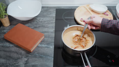 close-up of chef stirring a pot of soup on an electric stove in a modern kitchen, the sleek countertop, wooden cutting board, and open cookbook enhance the cozy, organized cooking environment