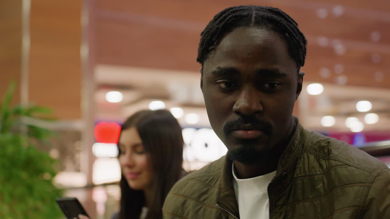Close up of sad man looking down with serious expression while smiling woman uses phone in background, set against colorful blurred lights and wooden decor inside bright modern shopping mall