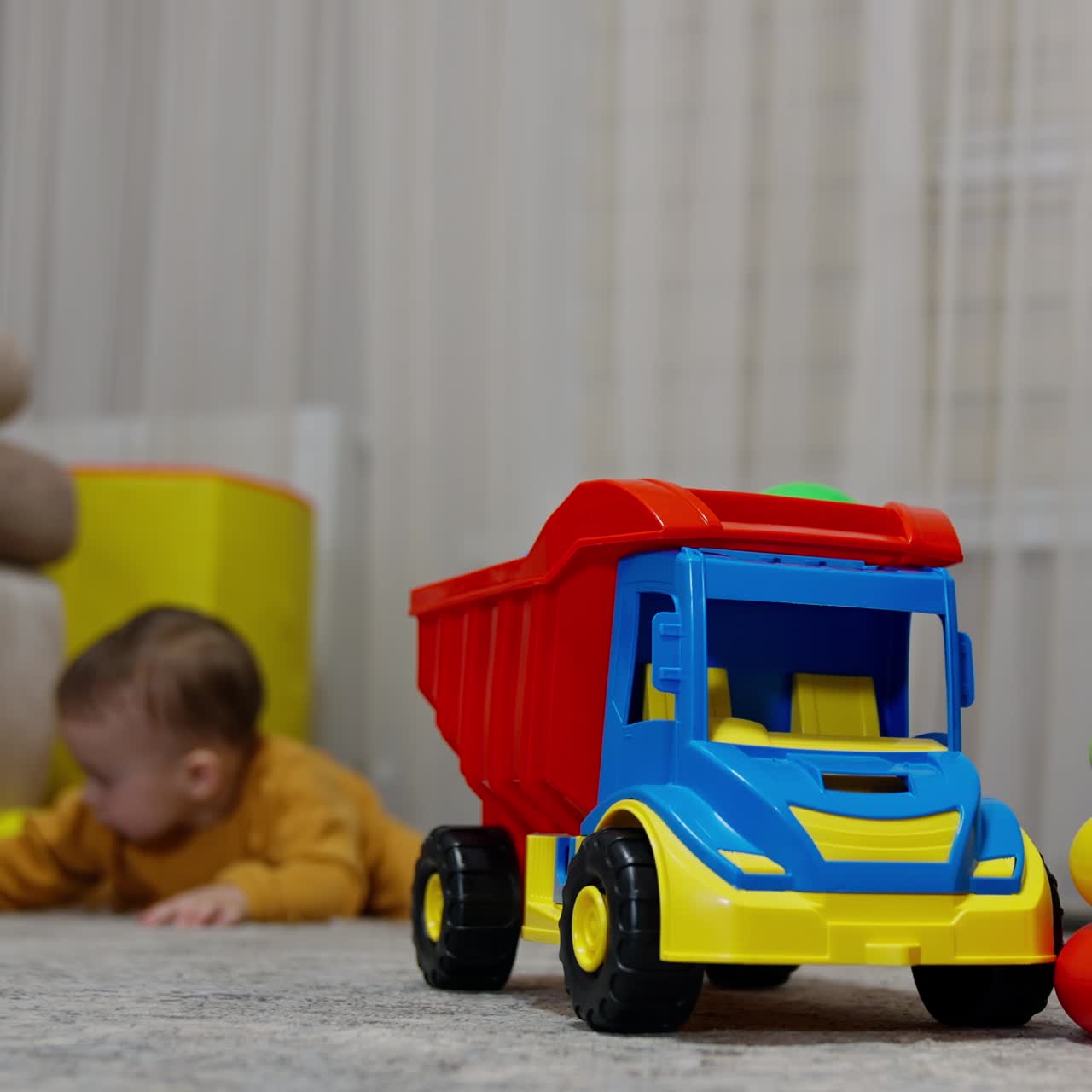 Toy lorry and pyramid on the floor in the room. Little kid at backdrop crawls up to play with toys. Blurred backdrop