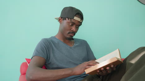 Close up of high school student in casual shirt and cap sitting on couch with crossed leg, opening novel to read as sunlight reflects on leg