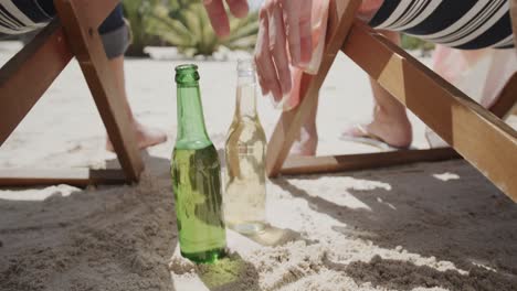 Happy-senior-caucasian-couple-sitting-in-deckchairs-on-beach-reaching-for-beers,-in-slow-motion