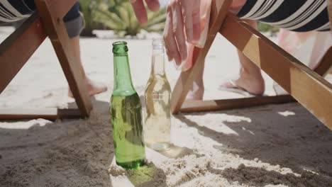una feliz pareja caucásica sentada en sillas de sol en la playa alcanzando cervezas, en cámara lenta