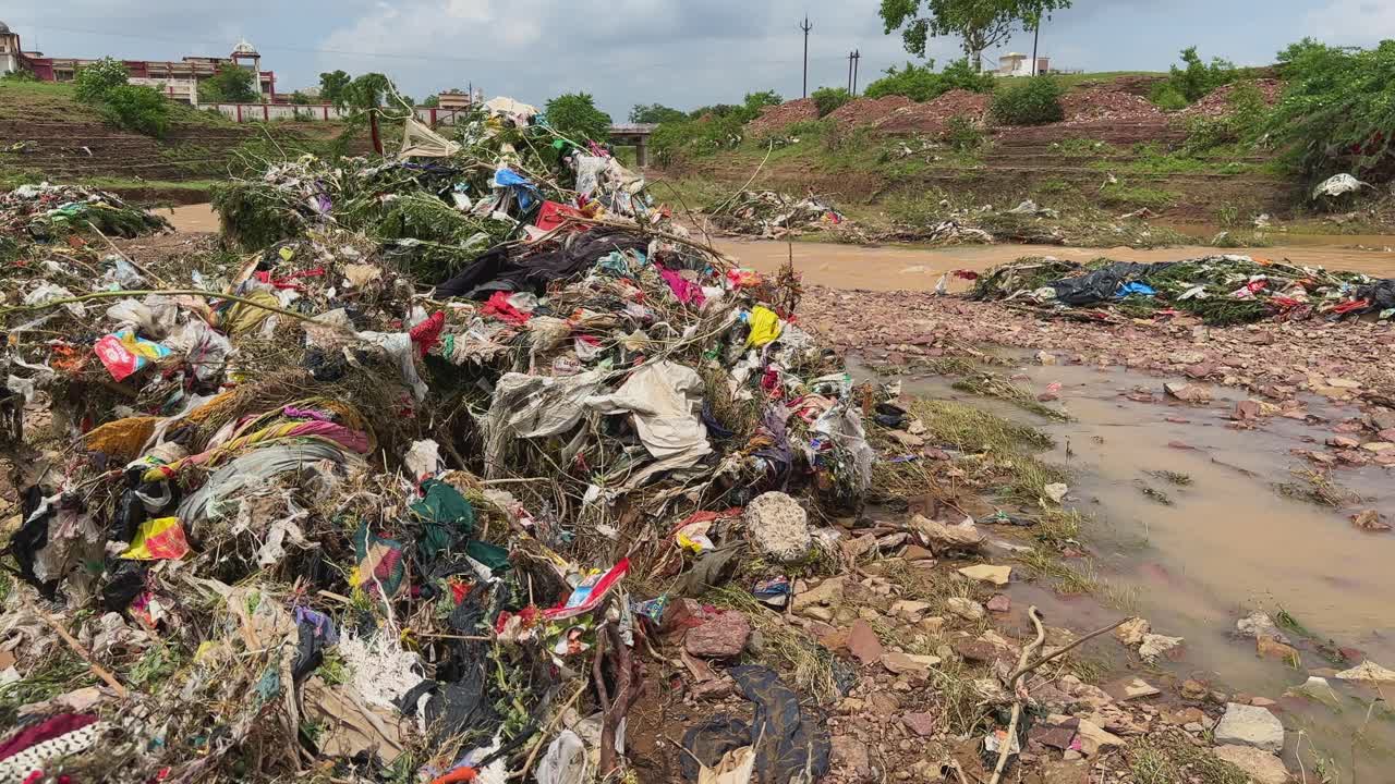 A heaps of garbage entangled in bushes and vegetation along the river floodplain, plastic bags, torn clothes, packaging materials, and organic waste carried and deposited by recent floodwaters