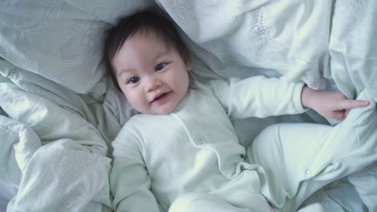아시안 아기 침대 위에 담요로 놀고 카메라를 바라보고 행복한 미소. 무고한 아이 미소 짓고 재미있는 순간을 즐기십시오. 아기 유아는 집에서 침실에서 재미있는 얼굴로 놀고 있습니다. 진정한 삶.
