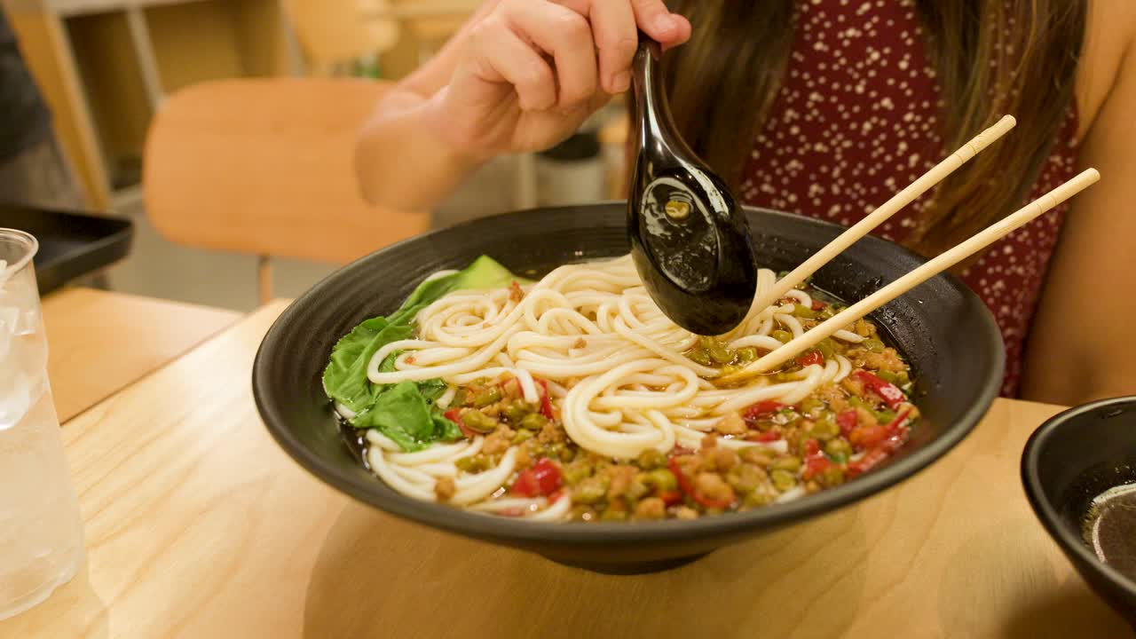 Young woman uses chopsticks and spoon to eat spicy noodle soup under warm indoor lighting
