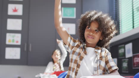 Verschiedene-Glückliche-Schulkinder-Sitzen-Am-Schreibtisch-Und-Heben-Die-Hände-Im-Klassenzimmer