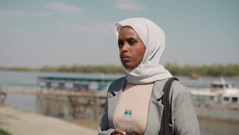 exhausted muslimah with bottle of water stands on riverbank