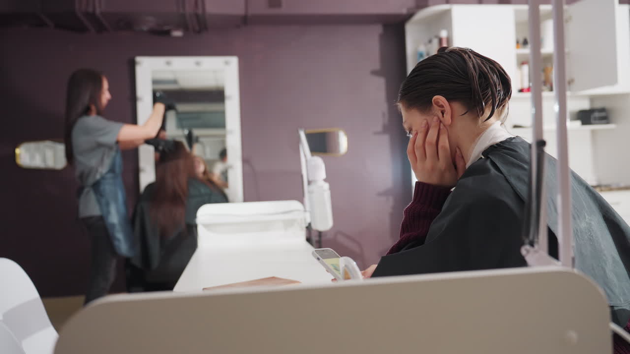 미용실 리셉션에서 불안하게 앉아 손으로 머리를 감싸고 휴대폰을 무릎에 둔 채 초조해하는 백인. 스타일리스트는 배경에서 다른 고객의 스타일링을 하고 있으며, 솔직하고 불안함과 걱정이 보이는 실내 장면입니다.