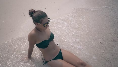 lady in orange sunglasses and black bikini sits on beach