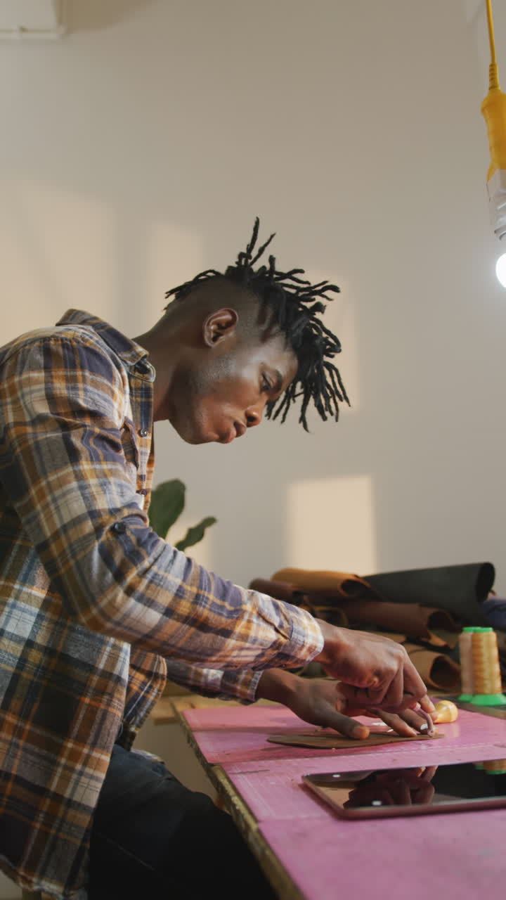 Vertical video of african american leather craftsman with dreadlocks using tool in workshop
