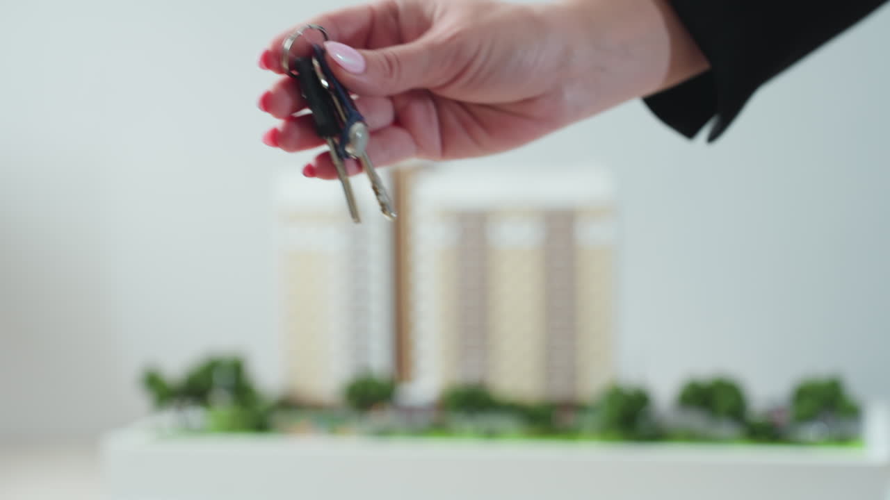 Close up of woman handing over key to another person, symbolizing successful real estate or business transaction, with background of blurred building model and indoor office environment