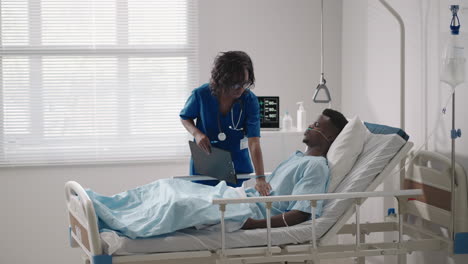 a black female doctor is talking to a black male patient lying on a hospital bed and connected to an acid supply