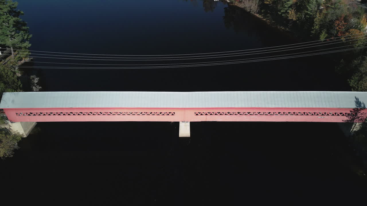 An aerial drone view tilting up to reveal a red and gray covered walking bridge across a river