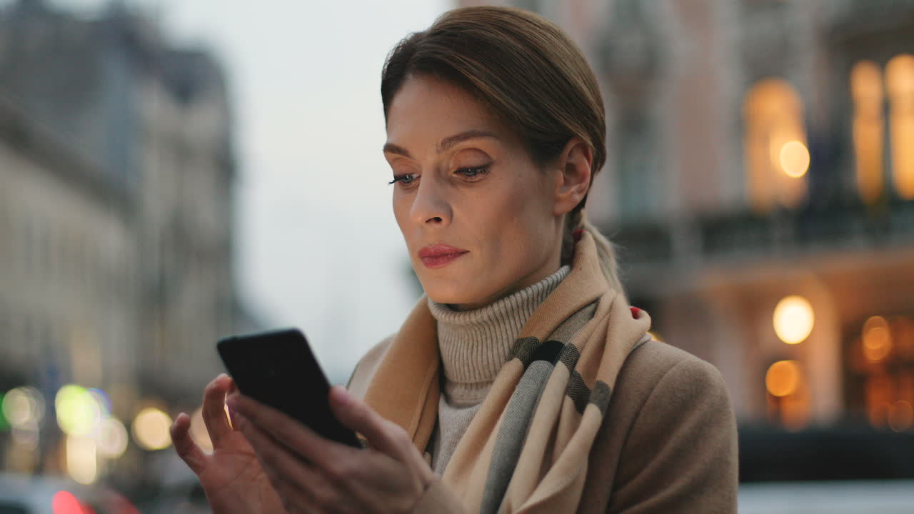 Close-up view of young Caucasian businesswoman texting on her smartphone in the street with city lights on the background